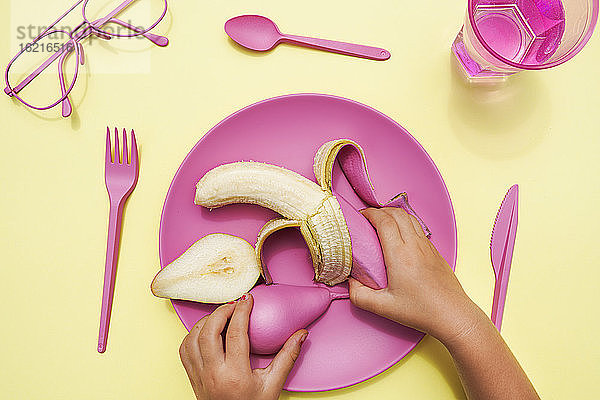 Hand eines kleinen Mädchens,  das eine pinkfarbene Birne und eine Banane von einem Plastikteller aufhebt