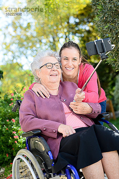 Fröhliche junge Frau macht ein Selfie mit einer älteren Frau in einem Garten eines Seniorenheims.