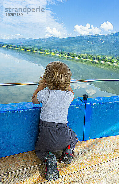 Kleiner Junge kniet vor einem Geländer und betrachtet die malerische Aussicht auf den Kerkini-See,  Mazedonien,  Griechenland