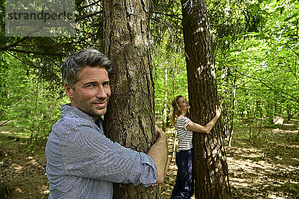 Lächelnder Mann und lächelnde Frau umarmen einen Baum,  während sie im Wald stehen