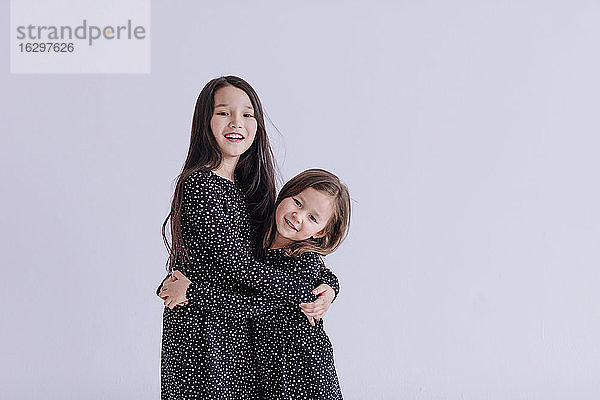 Lächelnde Schwestern,  die sich umarmen,  während sie vor einem weißen Hintergrund im Studio stehen