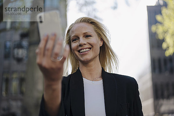 Lächelnde blonde weibliche Fachkraft bei einem Videoanruf über ein Smartphone in der Stadt