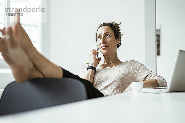 Geschäftsfrau mit Füßen auf dem Tisch,  die beim Entspannen über ein Smartphone spricht