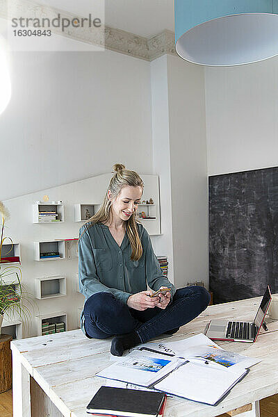 Lächelnde Frau sitzt auf dem Schreibtisch im Heimbüro und benutzt ein Smartphone
