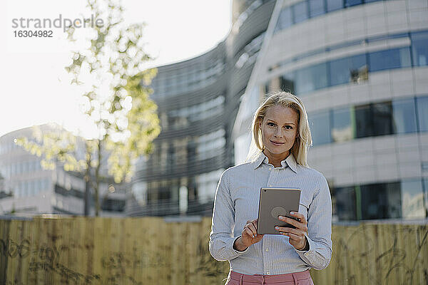 Selbstbewusste Geschäftsfrau,  die ein digitales Tablet hält,  während sie an einem Gebäude im Finanzviertel steht
