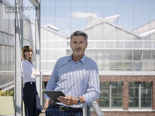 Selbstbewusster Geschäftsmann hält ein digitales Tablet,  während er mit einer Mitarbeiterin im Gewächshaus steht