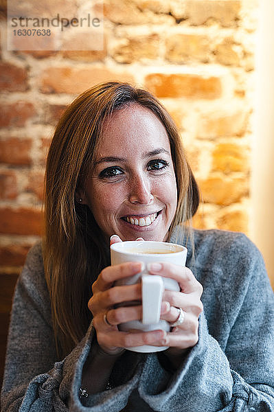 Frau lächelt,  während sie eine Kaffeetasse gegen eine Backsteinmauer in einem Kaffeehaus hält