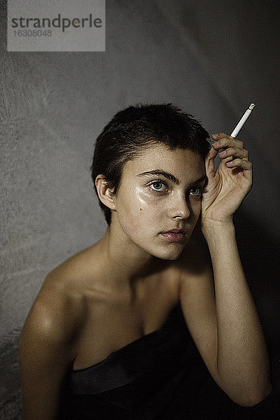Schöne junge Frau schaut weg,  während sie eine Zigarette gegen eine graue Wand hält