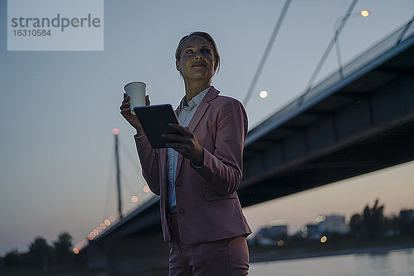 Geschäftsfrau schaut weg,  während sie ein digitales Tablet und einen Einwegbecher gegen eine Brücke in der Abenddämmerung hält