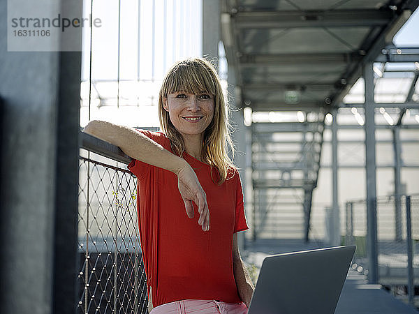 Lächelnde Geschäftsfrau,  die einen Laptop benutzt,  während sie an einem Geländer im Gewächshaus sitzt