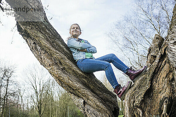 Lächelndes Mädchen mit verschränkten Armen an einen Baumstamm gelehnt im Park