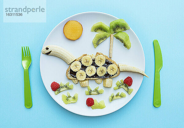 Frühstück mit Dinosaurierform aus Banane,  Kiwi,  Brot,  Schokoladenaufstrich und Beeren auf weißem Teller auf blauem Hintergrund