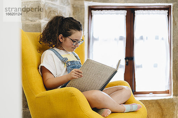 Mädchen liest ein Buch,  während sie zu Hause auf einem Sessel sitzt