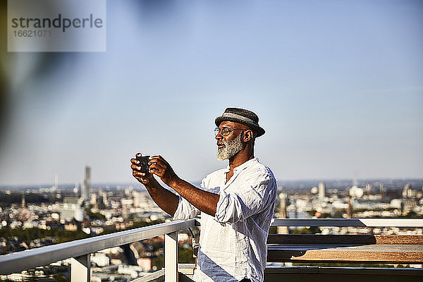 Älterer Mann,  der ein Selfie macht,  während er ein Smartphone benutzt,  das auf einer Gebäudeterrasse steht