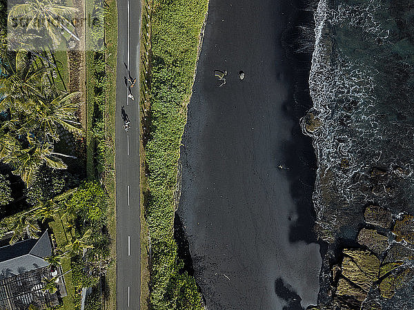 Indonesien,  Bali,  Luftaufnahme von Männern,  die auf einer asphaltierten Straße entlang eines schwarzen Küstenstrands Skateboard fahren