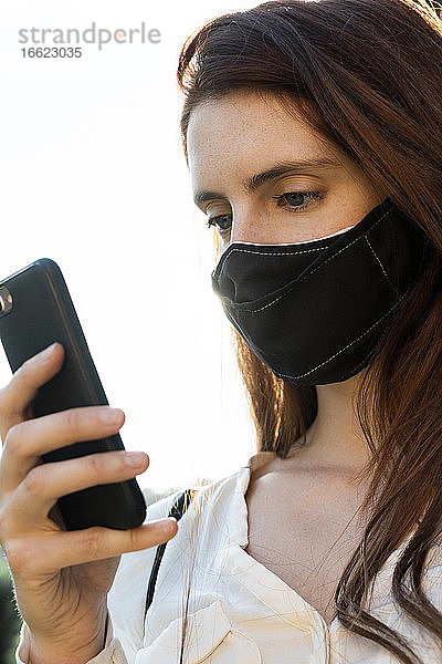 Junge Frau,  die eine Textnachricht auf ihrem Smartphone schreibt und eine Gesichtsmaske trägt