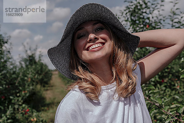 Nahaufnahme einer lächelnden erwachsenen Frau mit Hut in einem Obstgarten