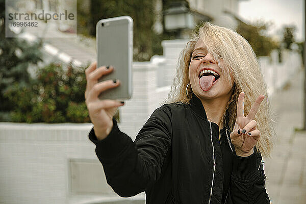 Glückliche junge Frau,  die ein Selfie macht,  während sie friedlich gestikuliert und die Zunge durch ihr Smartphone herausstreckt