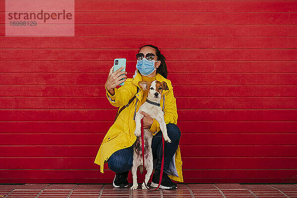 Frau mit Gesichtsmaske und Hund,  die Smartphone-Selfies vor einer roten Wand macht