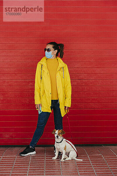 Frau mit Gesichtsmaske und Hund,  in gelbem Regenmantel vor roter Wand