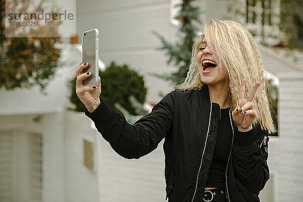 Blonde junge Frau macht Frieden Geste während der Einnahme selfie durch Handy