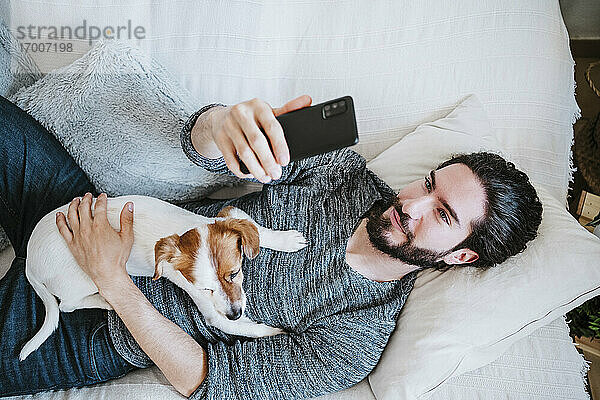 Junger Mann,  der ein Selfie mit seinem Handy macht,  während er mit seinem Hund zu Hause auf dem Sofa liegt