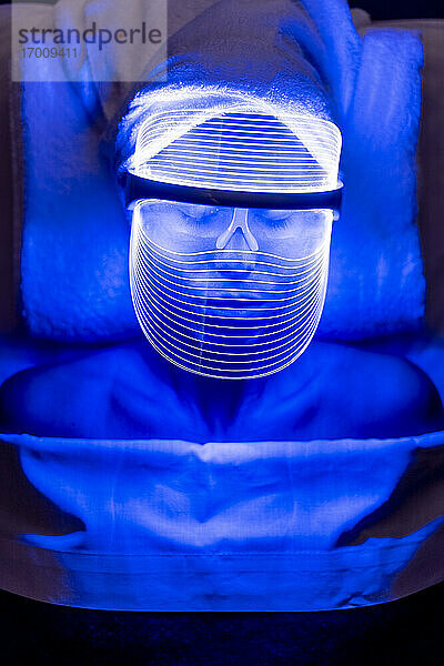 Junge Kundin trägt eine blau beleuchtete Maske während einer Verjüngungsbehandlung im Schönheitssalon