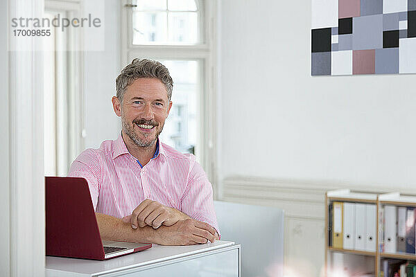 Lächelnde reife männliche Fachkraft mit Laptop am Schreibtisch in einer Bürokabine