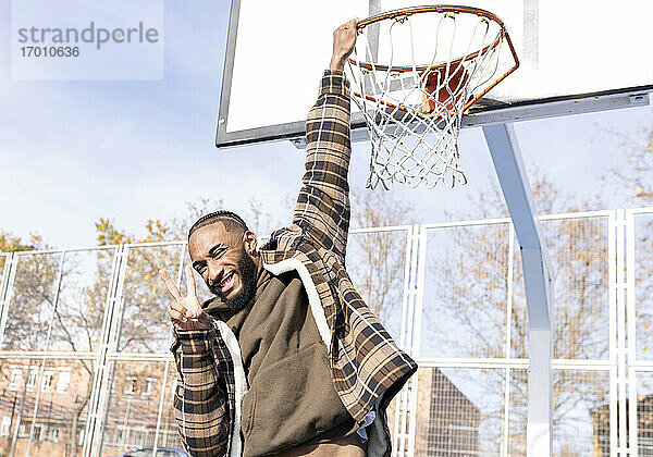 Glücklicher junger Mann,  der Frieden gestikuliert,  während er an einem sonnigen Tag am Basketballkorb hängt