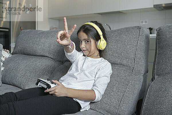 Kleines Mädchen macht Friedenszeichen,  während sie im Wohnzimmer ein Videospiel spielt