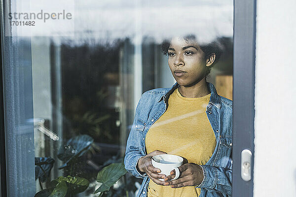 Frau schaut durch ein Fenster,  hält eine Kaffeetasse