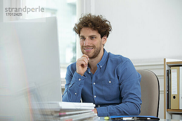 Lächelnder Geschäftsmann mit Hand am Kinn bei der Arbeit am Computer im Büro