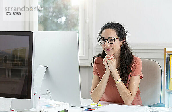 Lächelnde Geschäftsfrau mit Hand am Kinn bei der Arbeit in einer Bürokabine