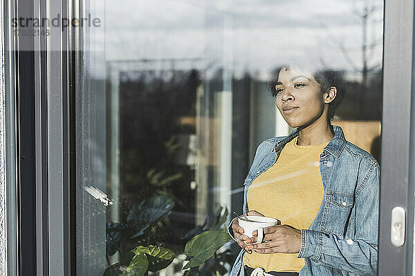 Frau schaut durch ein Fenster,  hält eine Kaffeetasse