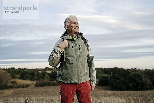 Lächelnder älterer männlicher Forscher auf einem landwirtschaftlichen Feld vor bewölktem Himmel am Wochenende