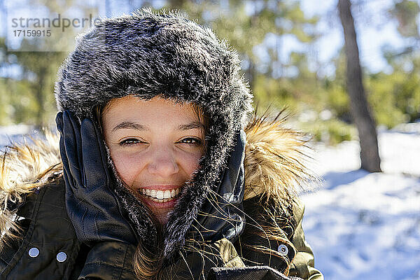 Fröhliche Frau mit Pelzmütze,  die im Winter lächelnd im Wald steht