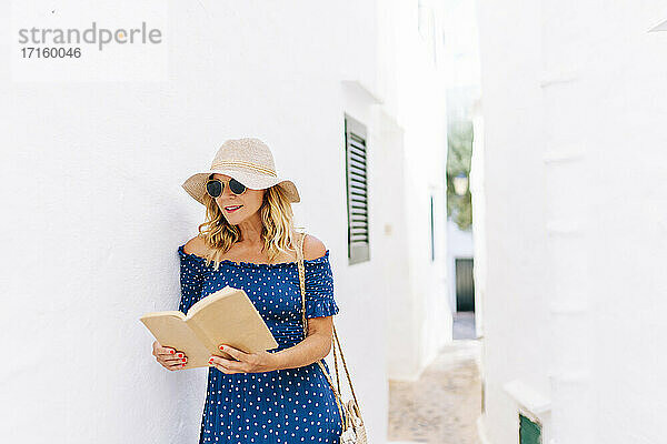 Frau mit Hut liest ein Buch,  während sie bei einem Haus im Dorf Binibeca,  Menorca,  Spanien,  steht