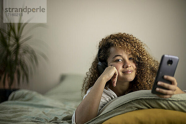 Lächelnde junge Frau mit Smartphone auf dem Bett im Schlafzimmer liegend