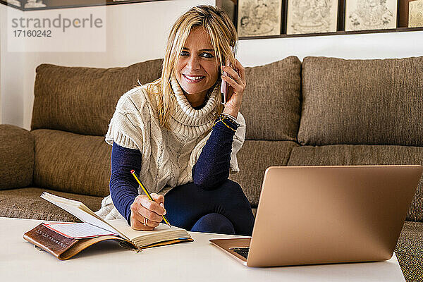 Lächelnde weibliche Therapeutin am Telefon mit Buch und Stift am Tisch am Arbeitsplatz sitzend