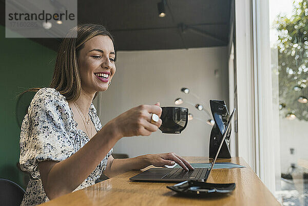 Glückliche Unternehmerin mit Kaffee in der Hand und Laptop auf dem Tisch im Café