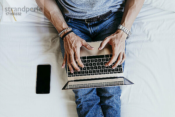 Draufsicht auf einen jungen Mann,  der zu Hause auf dem Bett sitzend an einem Laptop und einem Mobiltelefon arbeitet