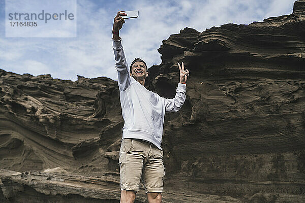 Lächelnder männlicher Tourist,  der Frieden gestikuliert,  während er ein Selfie vor einem Felsen in El golfo,  Lanzarote,  Spanien macht