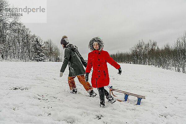 Teenager-Mädchen,  das einen Schlitten zieht,  während ein kleines Mädchen im Winter im Schnee läuft