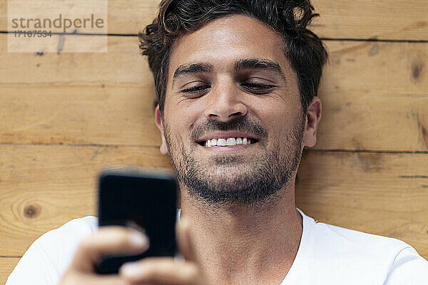 Lächelnder Mann,  der ein Mobiltelefon benutzt,  während er zu Hause auf dem Boden liegt