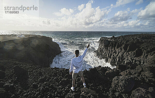 Junger männlicher Tourist,  der auf einem Felsen vor dem Meer in Los Hervideros,  Lanzarote,  Spanien,  steht und Frieden gestikuliert
