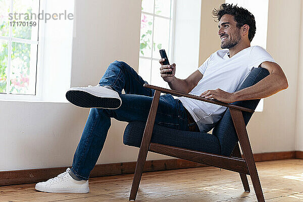 Lächelnder Mann,  der ein Smartphone benutzt,  während er sich zu Hause auf einem Sessel ausruht
