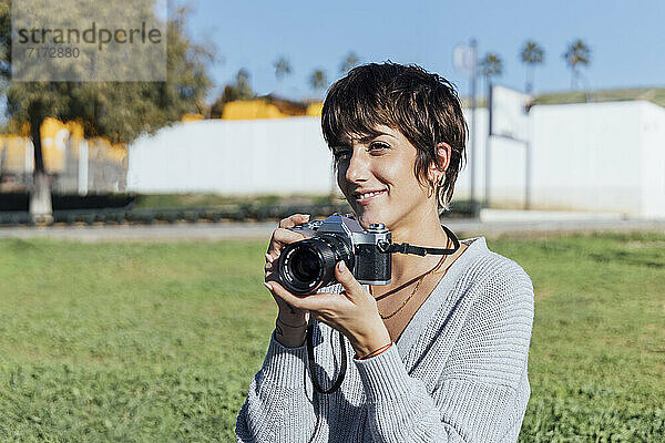 Lächelnde Frau mit analoger Kamera,  die im Park sitzt und lächelt