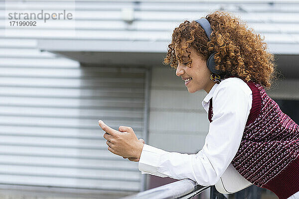 Lächelndes Teenager-Mädchen mit Kopfhörern,  das ein Mobiltelefon benutzt und sich auf ein Geländer stützt