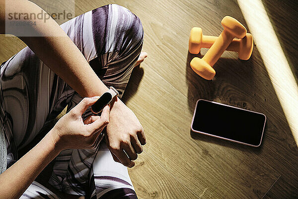 Junge Frau,  die eine intelligente Uhr benutzt,  während sie zu Hause im Schneidersitz auf dem Boden sitzt