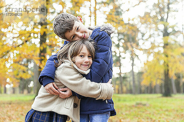 Lächelnder Bruder und lächelnde Schwester umarmen sich,  während sie im Wald stehen